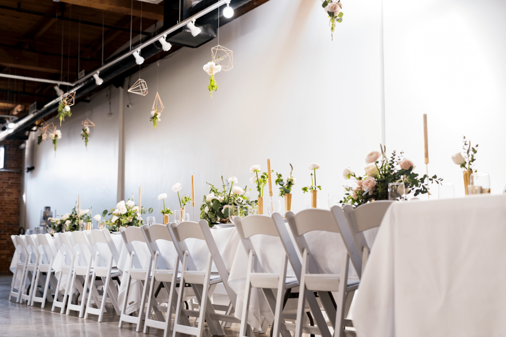 Long table draped in white table cloth and seated with white folding chairs sits below industrial lighting and decor hanging from the ceiling